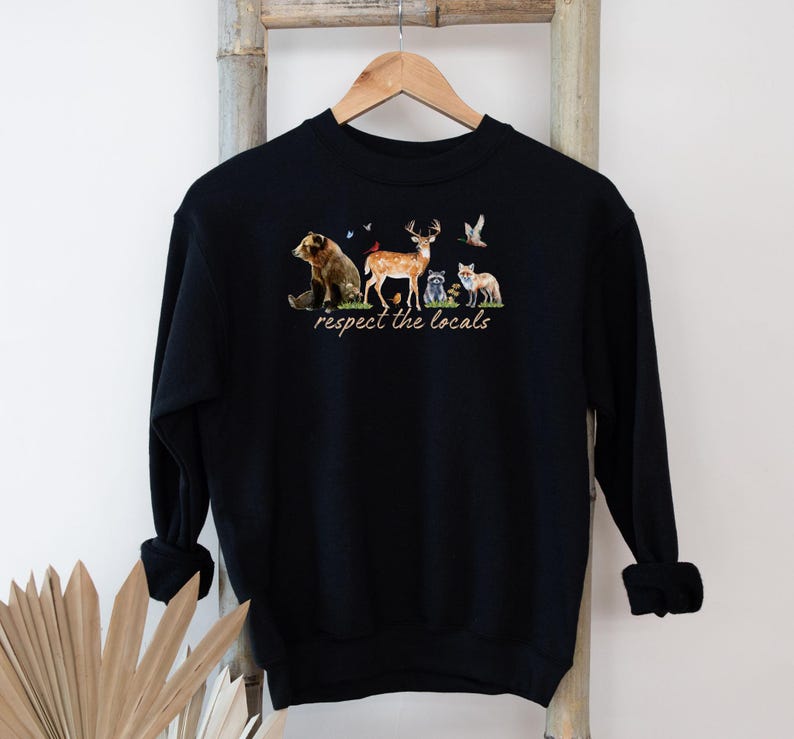 Op de afbeelding: Zwart sweatshirt met een aquarel illustratie van een beer, hert, wasbeer en vos. De tekst "respect the locals" is onder de dieren gedrukt. Het sweatshirt hangt aan een houten hanger.