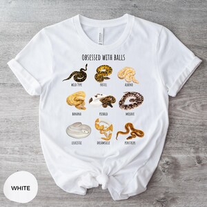 May include: White t-shirt with the text "OBSESSED WITH BALLS" and illustrations of various snake morphs, including Wild Type, Pastel, Albino, Banana, Piebald, Mojave, Leucistic, Dreamsicle, and Penstripe. The shirt is made of a soft material.