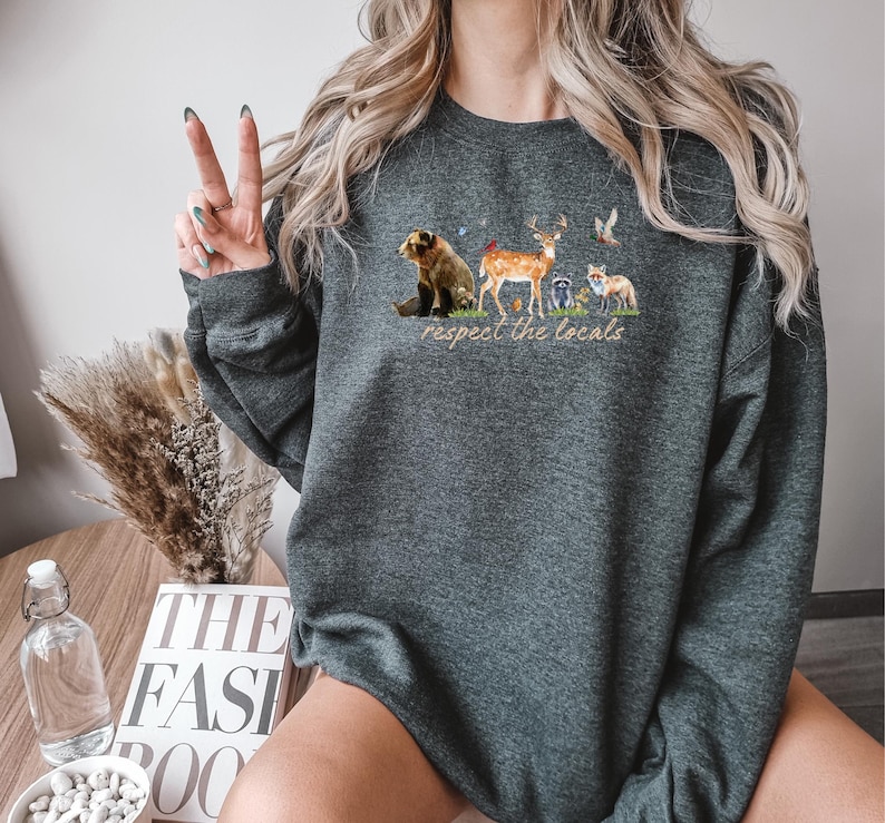 Op de afbeelding: Donkergrijze sweatshirt met een aquarelontwerp van een beer, hert, wasbeer en vos. De zin "respect the locals" staat onder de dieren gedrukt. De sweatshirt heeft een ronde hals.