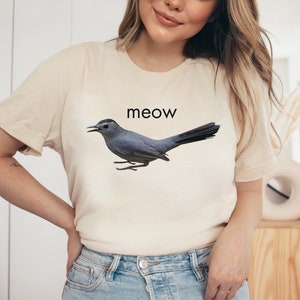 May include: A light beige t-shirt with a gray bird graphic and the word "meow" printed in black.