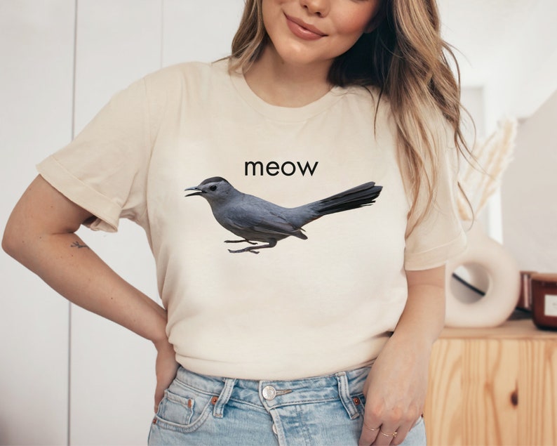 May include: A light beige t-shirt with a gray bird graphic and the word "meow" printed in black.