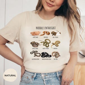 Puede incluir: Camiseta beige con la frase "NOODLE ENTHUSIAST" e ilustraciones de varias especies de serpientes, como la serpiente del ma&iacute;z, la boa de arena y la pit&oacute;n bola. La camiseta tambi&eacute;n presenta la palabra "NATURAL" en la parte inferior.
