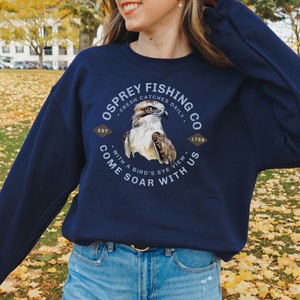 May include: Navy blue sweatshirt with a graphic of an osprey with the text "Osprey Fishing Co. Est. 1758 Fresh Catches Daily Come Soar With Us With a Bird's Eye View"