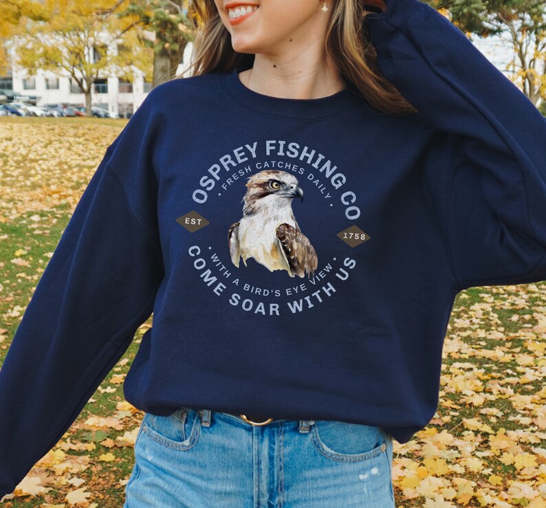 May include: Navy blue sweatshirt with a graphic of an osprey with the text "Osprey Fishing Co. Est. 1758 Fresh Catches Daily Come Soar With Us With a Bird's Eye View"