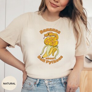 Op de afbeelding: Een wit T-shirt met een afbeelding van een gele banaan met een gele en bruine ball python eromheen gewikkeld. De tekst "Bananas about Ball Pythons" is in oranje rond de banaan en slang gedrukt.