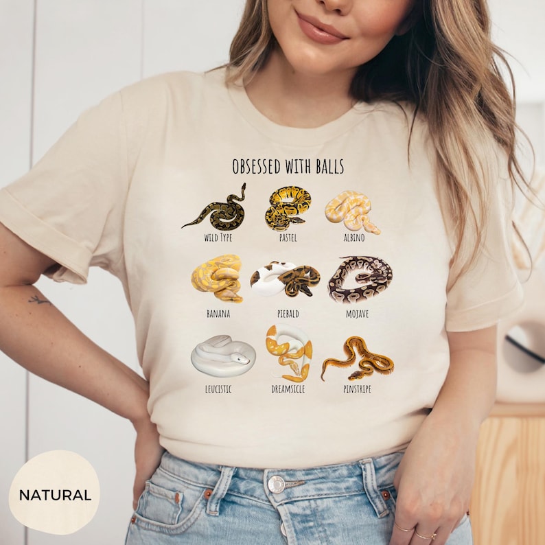 May include: A beige t-shirt with the text "OBSESSED WITH BALLS" and illustrations of various snake morphs, including Wild Type, Pastel, Albino, Banana, Piebald, Mojave, Leucistic, Dreamsicle, and Pinstripe. The shirt is labeled "NATURAL".