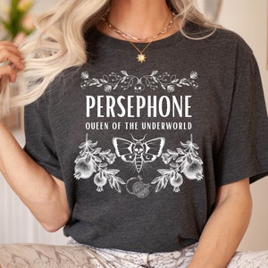 May include: A dark gray t-shirt with a white graphic design featuring the text "PERSEPHONE QUEEN OF THE UNDERWORLD" with a moth and floral accents.