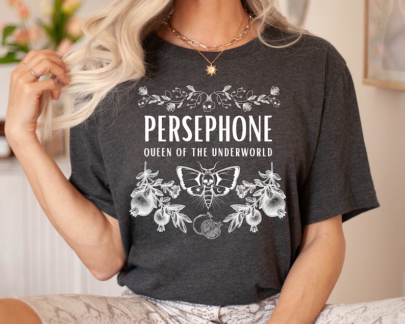 May include: A dark gray t-shirt with a white graphic design featuring the text "PERSEPHONE QUEEN OF THE UNDERWORLD" with a moth and floral accents.