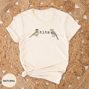 Puede incluir: Camiseta color crema con la palabra "tits" entre dos ilustraciones de p&aacute;jaros. Las aves est&aacute;n representadas de forma realista. La camiseta est&aacute; hecha de un material suave y c&oacute;modo, ideal para uso casual.