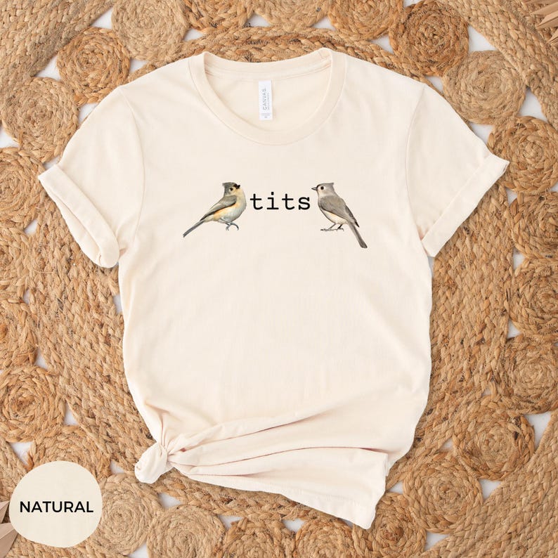Puede incluir: Camiseta color crema con la palabra "tits" entre dos ilustraciones de p&aacute;jaros. Las aves est&aacute;n representadas de forma realista. La camiseta est&aacute; hecha de un material suave y c&oacute;modo, ideal para uso casual.