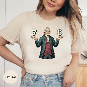 May include: A natural-colored t-shirt featuring a graphic of a man in a historical outfit holding the numbers 7 and 6. The man is wearing a green coat, a red vest, and a white ruffled shirt. The word "NATURAL" is printed at the bottom.