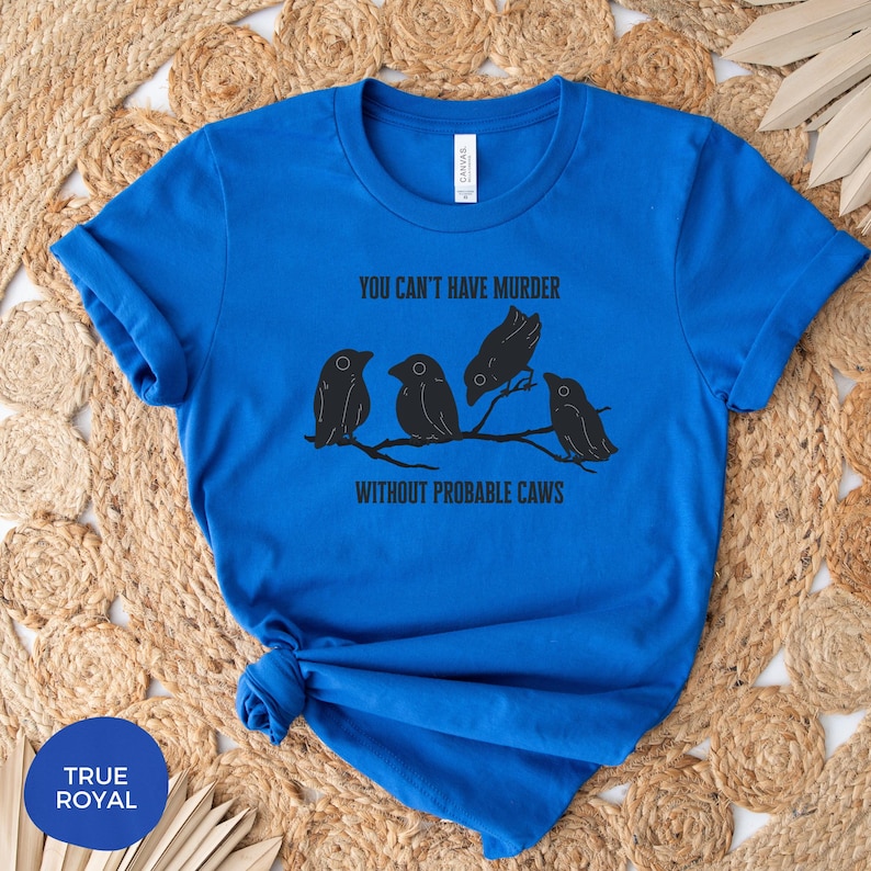 May include: Royal blue t-shirt with the text "YOU CAN'T HAVE MURDER WITHOUT PROBABLE CAWS" and a graphic of four black birds perched on a branch. The shirt is made of a soft material.