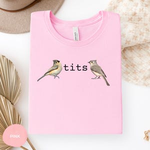 Puede incluir: Sudadera rosa claro con cuello redondo y la palabra "tits" en texto negro. Dos ilustraciones de p&aacute;jaros flanquean el texto. La sudadera est&aacute; doblada y presentada sobre una superficie blanca.