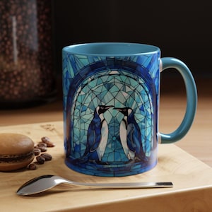 K&ouml;nnte beinhalten: Eine blaue Keramiktasse mit einem Buntglasdesign, das zwei Pinguine in einem blauen und wei&szlig;en Torbogen zeigt.