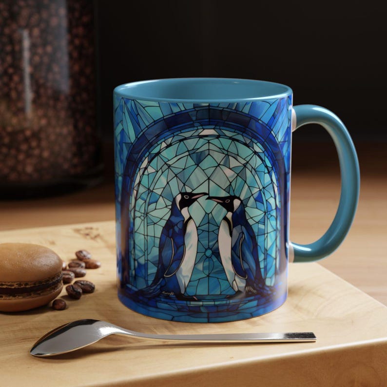 K&ouml;nnte beinhalten: Eine blaue Keramiktasse mit einem Buntglasdesign, das zwei Pinguine in einem blauen und wei&szlig;en Torbogen zeigt.