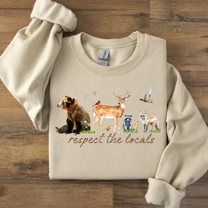 Op de afbeelding: Beige sweatshirt met een aquarel illustratie van bosdieren, waaronder een beer, een hert, een wasbeer en een vos. De tekst "respect the locals" is in bruin schrift gedrukt. De sweatshirt heeft een ronde hals en lange mouwen.