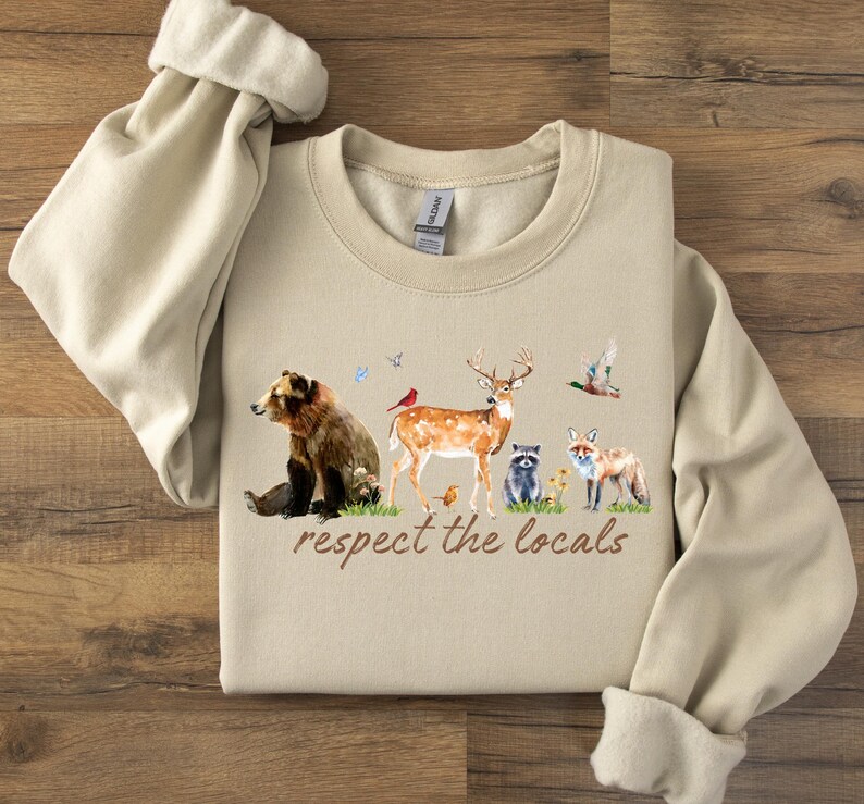 Op de afbeelding: Beige sweatshirt met een aquarel illustratie van bosdieren, waaronder een beer, een hert, een wasbeer en een vos. De tekst "respect the locals" is in bruin schrift gedrukt. De sweatshirt heeft een ronde hals en lange mouwen.