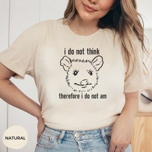 May include: A beige t-shirt with a black and white illustration of an opossum and the text "i do not think therefore i do not am".