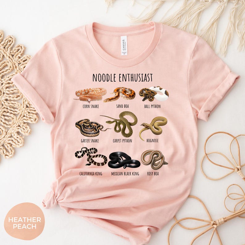 Puede incluir: Camiseta melocot&oacute;n con el texto "NOODLE ENTHUSIAST" e ilustraciones de varias especies de serpientes, como la Garter Snake y la Carpet Python. La camiseta es de material suave.