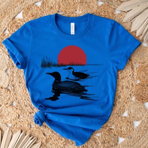 May include: A royal blue t-shirt with a black silhouette of two loons swimming in a lake with a red sun setting in the background.