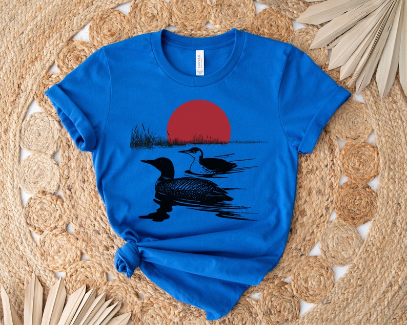 May include: A royal blue t-shirt with a black silhouette of two loons swimming in a lake with a red sun setting in the background.