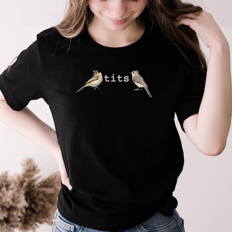 Puede incluir: Camiseta negra con dos ilustraciones de p&aacute;jaros y la palabra "tits" impresa entre ellas. Los p&aacute;jaros son en tonos marrones, grises y amarillos. La camiseta es de cuello redondo.