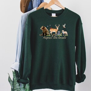 Op de afbeelding: Een donkergroene sweatshirt met een afbeelding van bosdieren en de tekst "respect the locals". Het ontwerp omvat een beer, een hert, een wasbeer en een vos. De sweatshirt hangt aan een houten hanger.