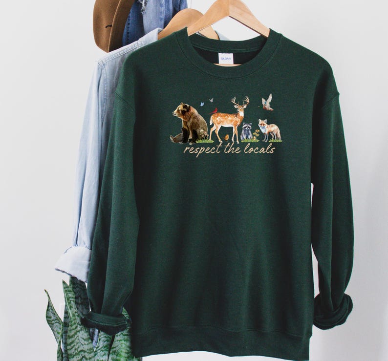 Op de afbeelding: Een donkergroene sweatshirt met een afbeelding van bosdieren en de tekst "respect the locals". Het ontwerp omvat een beer, een hert, een wasbeer en een vos. De sweatshirt hangt aan een houten hanger.