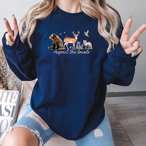 Op de afbeelding: Marineblauwe sweatshirt met een aquarel illustratie van bosdieren, waaronder een beer, een hert, een wasbeer en een vos. De tekst "respect the locals" is eronder gedrukt. Het sweatshirt heeft een casual stijl.
