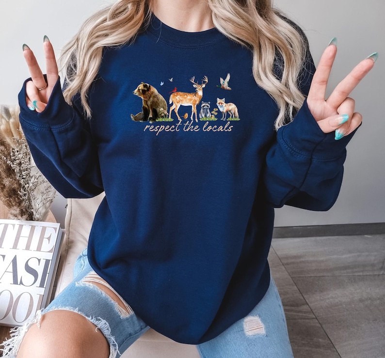 Op de afbeelding: Marineblauwe sweatshirt met een aquarel illustratie van bosdieren, waaronder een beer, een hert, een wasbeer en een vos. De tekst "respect the locals" is eronder gedrukt. Het sweatshirt heeft een casual stijl.