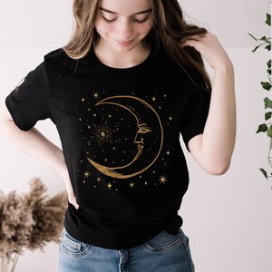 Pode incluir: Uma camiseta preta com um design de lua crescente dourada e estrelas. A lua tem um rosto e &eacute; cercada por pequenas estrelas.