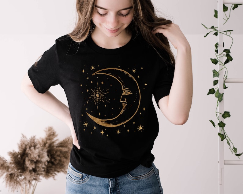 Pode incluir: Uma camiseta preta com um design de lua crescente dourada e estrelas. A lua tem um rosto e &eacute; cercada por pequenas estrelas.