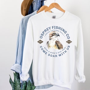 May include: White sweatshirt with a blue and brown graphic design of an osprey with the text "Osprey Fishing Co. Fresh Catches Daily. Est. 1758. Come Soar With Us. With a Bird's Eye View."