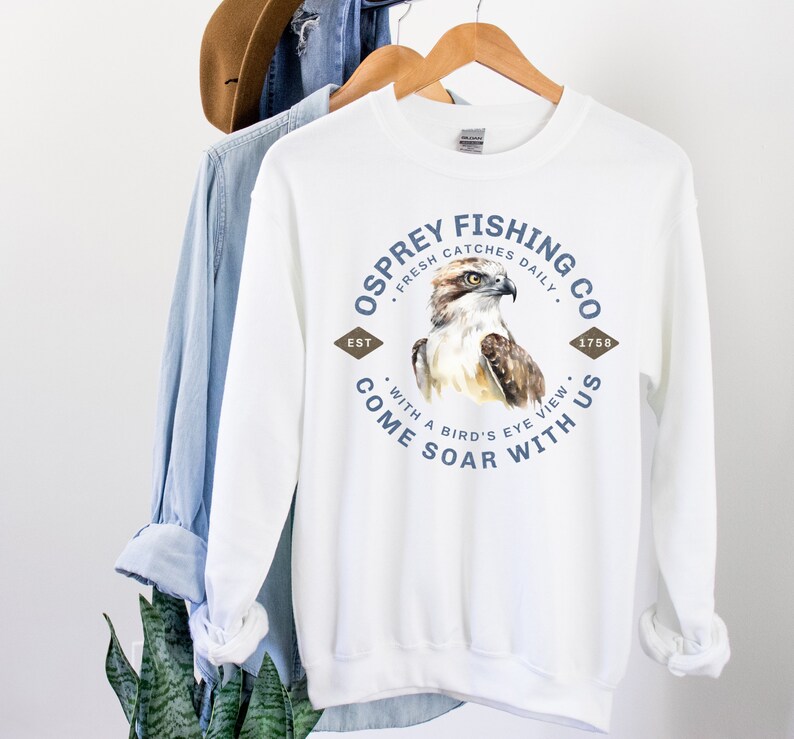 May include: White sweatshirt with a blue and brown graphic design of an osprey with the text "Osprey Fishing Co. Fresh Catches Daily. Est. 1758. Come Soar With Us. With a Bird's Eye View."