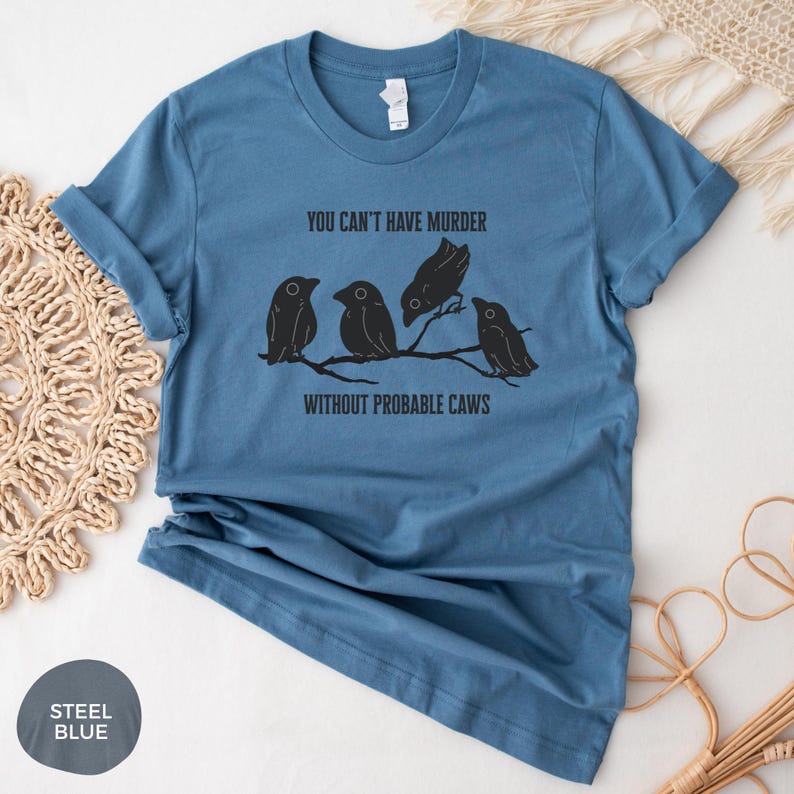 May include: Steel blue t-shirt with the text "YOU CAN'T HAVE MURDER WITHOUT PROBABLE CAWS" and a graphic of four black birds perched on a branch. The shirt has rolled-up sleeves.