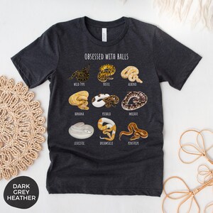 May include: Dark grey heather t-shirt with the text "OBSESSED WITH BALLS" and illustrations of various ball python morphs, including Wild Type, Pastel, Albino, Banana, Piebald, Mojave, Leucistic, Dreamsicle, and Pinstripe.