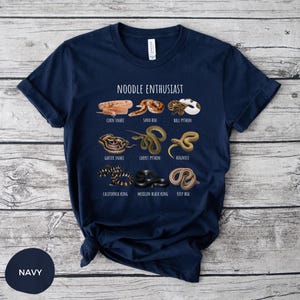 Puede incluir: Camiseta azul marino con ilustraciones de varias especies de serpientes, incluyendo serpiente del ma&iacute;z y pit&oacute;n bola, con el texto "NOODLE ENTHUSIAST" encima. La camiseta est&aacute; hecha de un material suave.