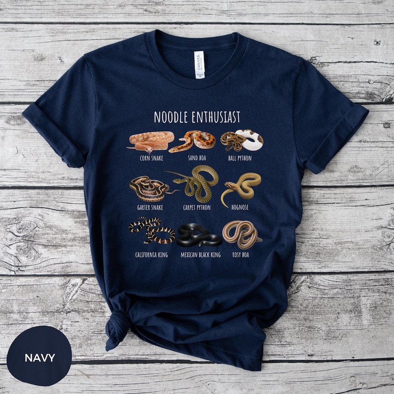 Puede incluir: Camiseta azul marino con ilustraciones de varias especies de serpientes, incluyendo serpiente del ma&iacute;z y pit&oacute;n bola, con el texto "NOODLE ENTHUSIAST" encima. La camiseta est&aacute; hecha de un material suave.