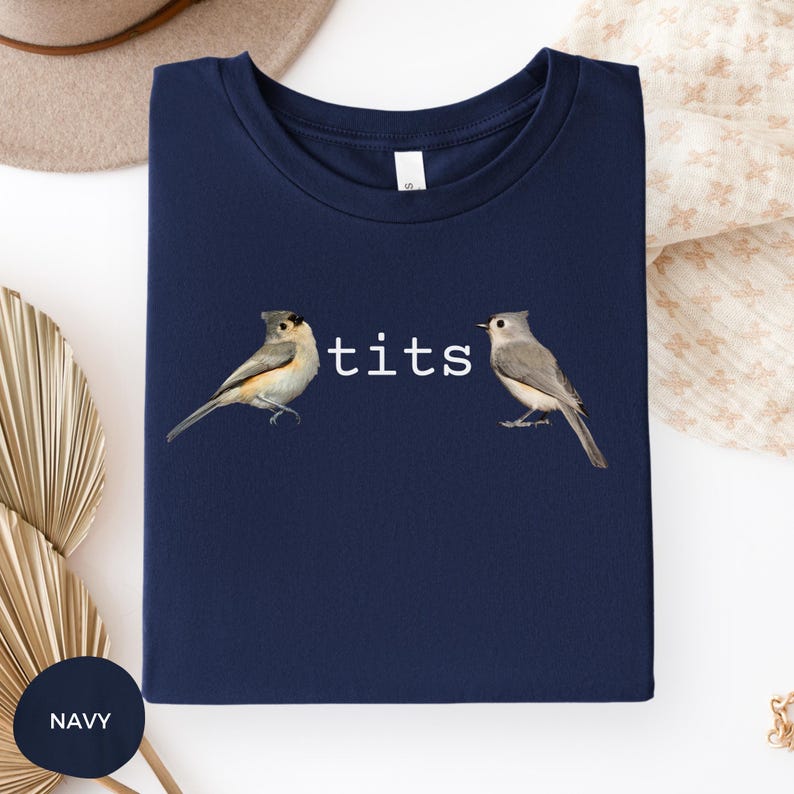 Puede incluir: Camiseta azul marino con la palabra "tits" en blanco entre dos ilustraciones de p&aacute;jaros. Los p&aacute;jaros son en tonos grises, marrones y blancos. La camiseta es de color azul marino liso.