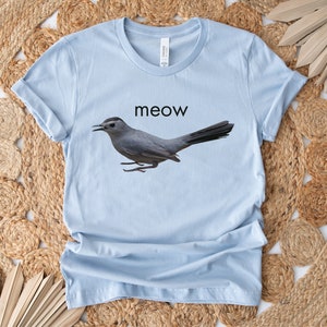 May include: Light blue t-shirt with a gray bird graphic and the word "meow" printed in black.