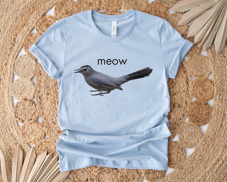 May include: Light blue t-shirt with a gray bird graphic and the word "meow" printed in black.