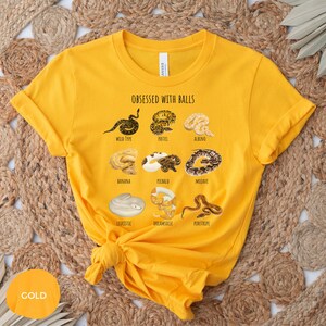May include: Mustard yellow t-shirt with the text "OBSESSED WITH BALLS" and illustrations of various snake morphs, including Wild Type, Pastel, Albino, Banana, Piebald, Mojave, Leucistic, Dreamsicle, and Pinstripe. The shirt has a crew neck and short sleeves.