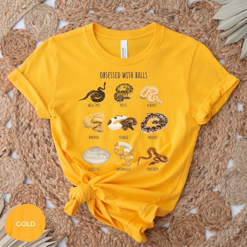 May include: Mustard yellow t-shirt with the text "OBSESSED WITH BALLS" and illustrations of various snake morphs, including Wild Type, Pastel, Albino, Banana, Piebald, Mojave, Leucistic, Dreamsicle, and Pinstripe. The shirt has a crew neck and short sleeves.