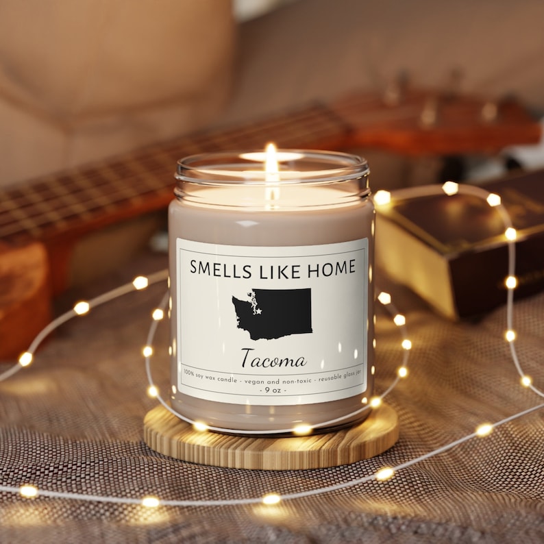 May include: A brown soy wax candle in a clear glass jar with a white label that reads "SMELLS LIKE HOME" and features a black silhouette of the state of Washington with the word "Tacoma" underneath. The candle is on a wooden coaster and surrounded by white string lights.