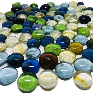 May include: A collection of smooth, round glass pebbles in various colors, including blue, green, clear, and brown. The pebbles are scattered on a white surface.