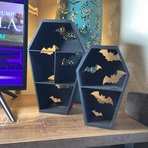 May include: Two black coffin-shaped shelves with gold bat cutouts. The shelves have multiple compartments and are made of wood. The shelves are on a wooden surface, and a teal decorative lantern is in the background. The shelves are a Halloween decoration.