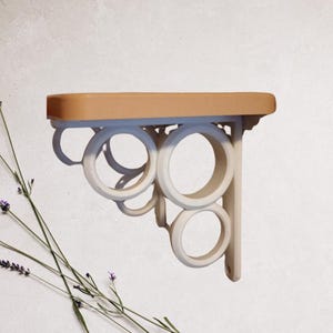 May include: A white shelf bracket with a decorative design of interlocking circles. The shelf is made of wood and is painted a light brown color.