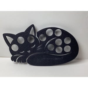 May include: Black cat-shaped organizer with multiple circular holes. The cat is in a sleeping position, with its paws tucked in. The organizer is made of a textured material and is designed to hold small items.