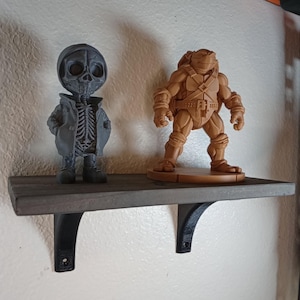 May include: A gray wooden shelf with black metal brackets holds two figurines. One is a gray skeleton figure wearing a coat. The other is a tan Teenage Mutant Ninja Turtle figurine. The shelf is mounted on a white wall.