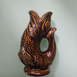 May include: A glossy brown ceramic pitcher shaped like a fish with its mouth open at the top. The body features detailed scale textures, a prominent eye, and a curved tail that forms a handle. The base is circular and supports the upright fish figure.